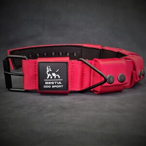May include: A bright red dog collar featuring a black metal buckle and a triangular D-ring. A black rectangular patch on the front displays a white dog silhouette logo and the text BESTIA DOG SPORT. The collar has a black interior lining.