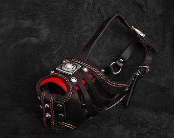 May include: Black leather dog muzzle with red stitching and a silver metal accent. The muzzle has a strap that goes around the dog's head.