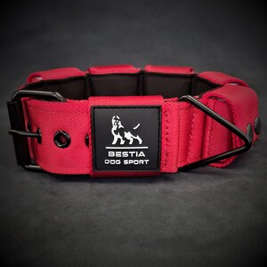 May include: A wide red dog collar featuring a black interior lining, black metal hardware, and a black rectangular patch on the front. The patch displays a white dog silhouette graphic above the text BESTIA DOG SPORT in white capital letters.