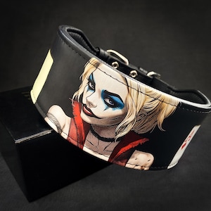 May include: Black leather dog collar featuring a colourful illustration of Harley Quinn from the DC Comics universe. The collar has a silver buckle and stitching.