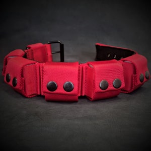 May include: A bright red utility belt featuring multiple small, rectangular pouches attached along the length of the strap. Each pouch is secured with two dark metal snap buttons. The belt includes a black metal buckle and a black interior lining.