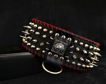 Puede incluir: Un collar de perro de cuero negro con tachuelas plateadas y costuras rojas. El collar tiene una hebilla de metal decorativa con un diseño.