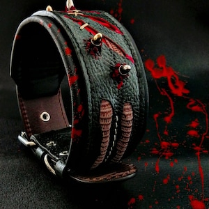 May include: A black leather dog collar with red paint splatters and silver studs. The collar has a brown leather lining and a silver buckle.
