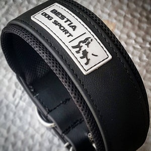 Puede incluir: Collar de perro de cuero negro con forro de malla blanca y un parche rectangular blanco que dice "BESTIA DOG SPORT" con una silueta de perro estilizada.
