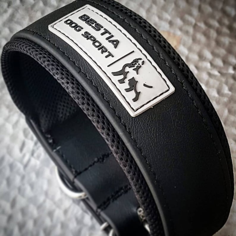 Puede incluir: Collar de perro de cuero negro con forro de malla blanca y un parche rectangular blanco que dice "BESTIA DOG SPORT" con una silueta de perro estilizada.