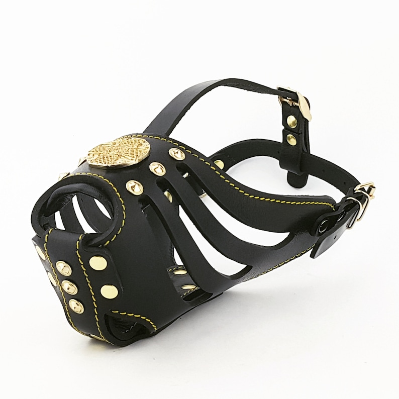May include: Black leather dog muzzle with gold accents and a decorative heart-shaped plate. The muzzle has a strap with a buckle for adjustment.