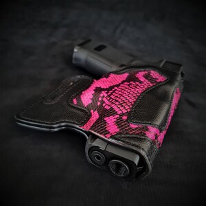 Könnte beinhalten: Ein schwarzes Lederholster mit einem rosafarbenen Schlangenhautmuster. Das Holster ist für eine Pistole konzipiert und hat einen Ausschnitt für den Lauf.