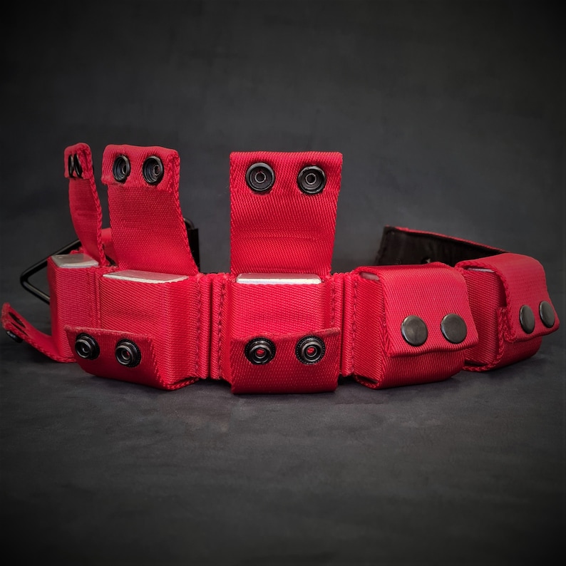 May include: A bright red nylon utility belt featuring multiple integrated pouches with black metal snap button closures. The belt includes several vertical flaps that secure over the pouches, with a black buckle visible on the left side.