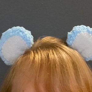 May include: A pair of light blue fuzzy ear headbands with white centers. The headbands are worn on a person's head.