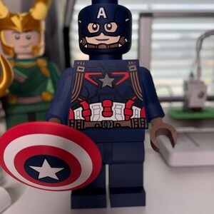 May include: A Captain America action figure in a block style, holding a red, white, and blue shield with a white star. The figure is dark blue with red and white accents and a helmet with the letter "A". Another figure is visible in the background.