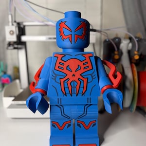 Op de afbeelding: Een grote blauwe figuur die lijkt op een Lego-figuur, met rode accenten. De figuur heeft een gedetailleerd ontwerp op het hoofd, de borst en de benen, met rode armbevestigingen. Het algehele ontwerp doet denken aan een superheld.