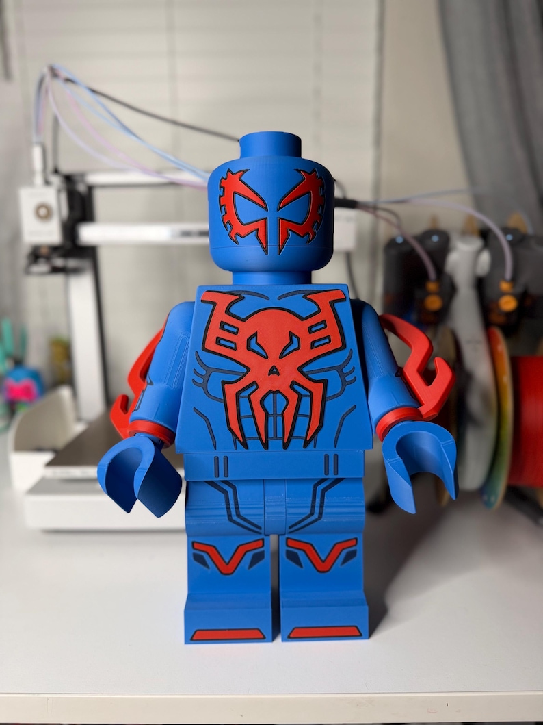 Op de afbeelding: Een grote blauwe figuur die lijkt op een Lego-figuur, met rode accenten. De figuur heeft een gedetailleerd ontwerp op het hoofd, de borst en de benen, met rode armbevestigingen. Het algehele ontwerp doet denken aan een superheld.
