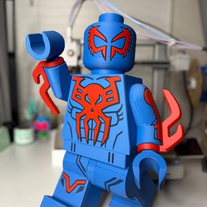 Op de afbeelding: Een blauw en rood actiefiguur in de stijl van een populair bouwsteenspeelgoed. De figuur heeft een blauw lichaam met rode accenten, waaronder een masker, borstembleem en armbevestigingen. De figuur poseert met &eacute;&eacute;n arm omhoog.