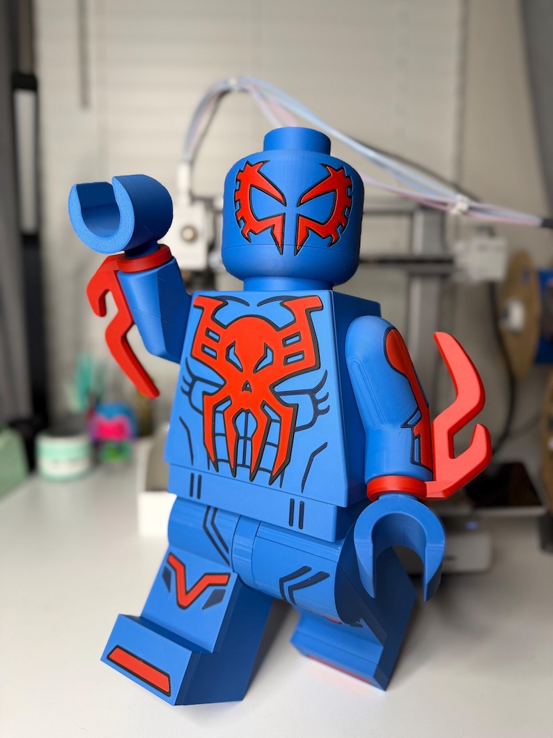 Op de afbeelding: Een blauw en rood actiefiguur in de stijl van een populair bouwsteenspeelgoed. De figuur heeft een blauw lichaam met rode accenten, waaronder een masker, borstembleem en armbevestigingen. De figuur poseert met &eacute;&eacute;n arm omhoog.