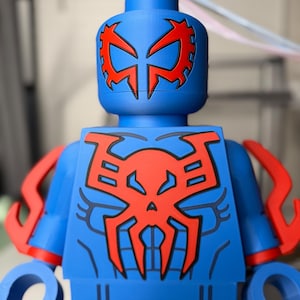 Op de afbeelding: Een blauw actiefiguur met rode accenten, die op een superheld lijkt. Het figuur heeft een rood spinnenembleem op de borst en het hoofd, met rode armdetails. Het figuur is gemaakt van plastic en heeft een blokkerig ontwerp.