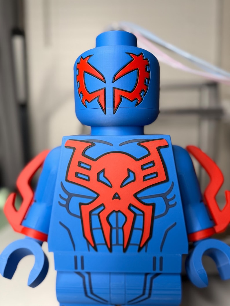 Op de afbeelding: Een blauw actiefiguur met rode accenten, die op een superheld lijkt. Het figuur heeft een rood spinnenembleem op de borst en het hoofd, met rode armdetails. Het figuur is gemaakt van plastic en heeft een blokkerig ontwerp.