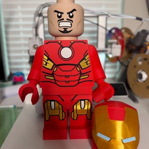 May include: A large red and gold Iron Man Lego figure with a removable helmet. The figure has a detailed red and gold suit design, a tan head with a stern expression, and is approximately 12 inches tall.