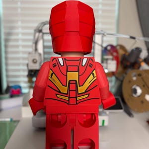 May include: A red Lego-style figure, reminiscent of Iron Man, viewed from the back. The figure has a red helmet and body with gold and black accents on the back. The figure is standing on a white surface.