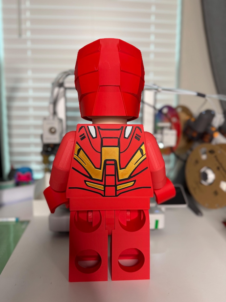 May include: A red Lego-style figure, reminiscent of Iron Man, viewed from the back. The figure has a red helmet and body with gold and black accents on the back. The figure is standing on a white surface.