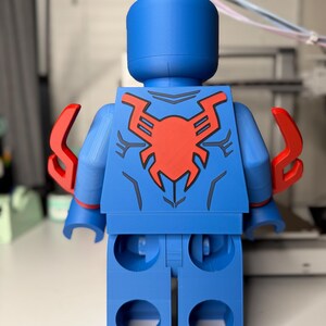 Op de afbeelding: Een grote blauwe figuur die lijkt op een klassieke speelfiguur, met een rood spinnenembleem op de rug. De figuur heeft rode accenten op de armen en een cilindrische kop. Het ontwerp doet denken aan een superheld.