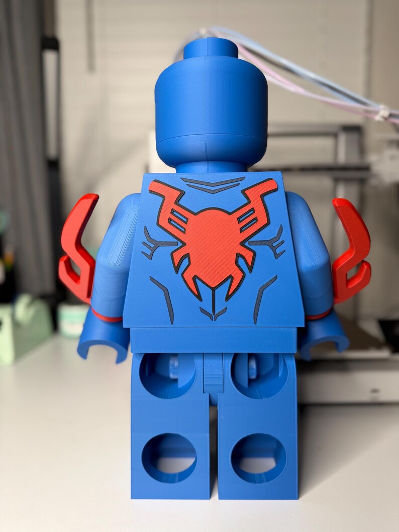 Op de afbeelding: Een grote blauwe figuur die lijkt op een klassieke speelfiguur, met een rood spinnenembleem op de rug. De figuur heeft rode accenten op de armen en een cilindrische kop. Het ontwerp doet denken aan een superheld.
