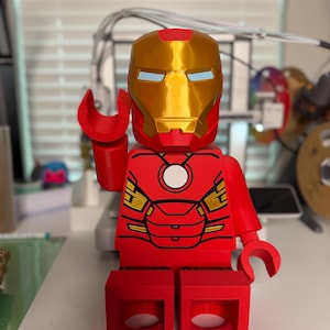 May include: A red and gold Iron Man figure in a Lego style. The figure has a gold helmet with blue eye slits, red arms and legs, and a red torso with gold accents. The figure is posed with one hand raised.