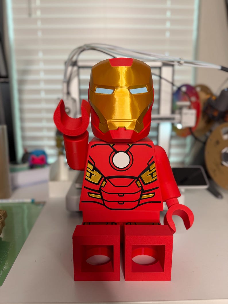 May include: A red and gold Iron Man figure in a Lego style. The figure has a gold helmet with blue eye slits, red arms and legs, and a red torso with gold accents. The figure is posed with one hand raised.