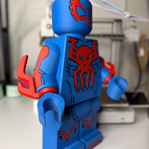 Op de afbeelding: Een grote blauwe figuur die lijkt op een Lego-minifiguur, met rode accenten en een gedetailleerd ontwerp. De figuur heeft een tandwielachtig embleem op het hoofd en een spinnenachtig embleem op de borst. De armen en benen hebben rode details.