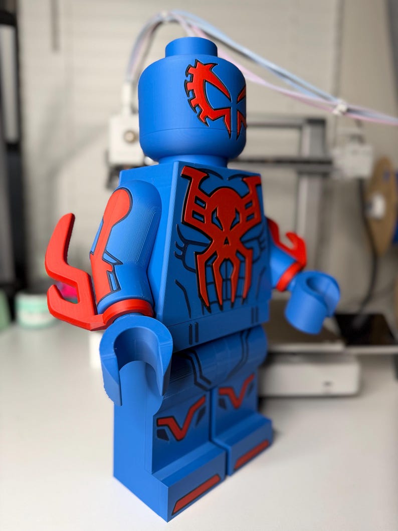 Op de afbeelding: Een grote blauwe figuur die lijkt op een Lego-minifiguur, met rode accenten en een gedetailleerd ontwerp. De figuur heeft een tandwielachtig embleem op het hoofd en een spinnenachtig embleem op de borst. De armen en benen hebben rode details.