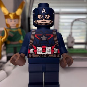 May include: A Captain America Lego figure, primarily navy blue, with a white "A" on the helmet. The figure has a star on the chest and brown hands. Another Lego figure, Loki, is visible in the background.