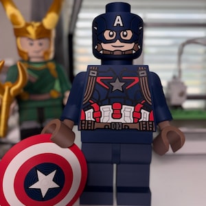 May include: A Captain America Lego figure, featuring a blue and red costume with a star emblem and a matching shield. The figure has a blue helmet with the letter "A" and brown arms. Another Lego figure of Loki is visible in the background.