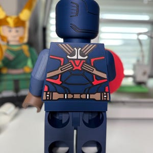 May include: A large, navy blue Captain America-themed Lego figure. The figure has a detailed design on the back with red, brown, and white accents. The figure is standing upright, with a red shield visible on the right side.