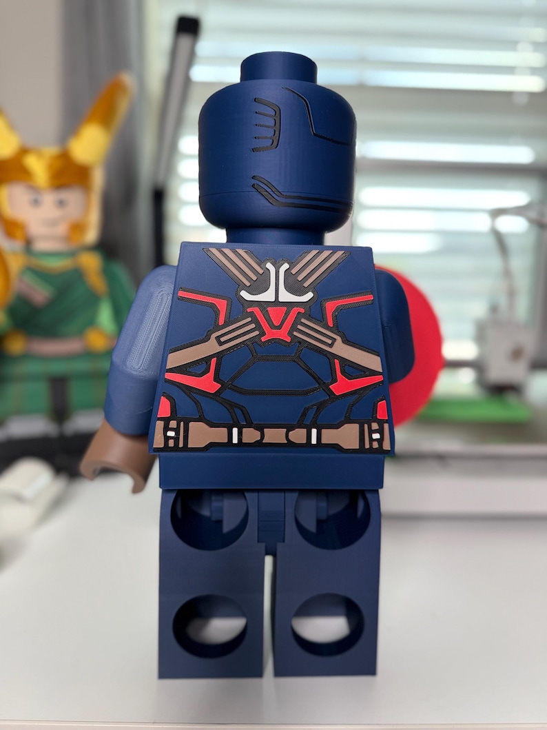 May include: A large, navy blue Captain America-themed Lego figure. The figure has a detailed design on the back with red, brown, and white accents. The figure is standing upright, with a red shield visible on the right side.