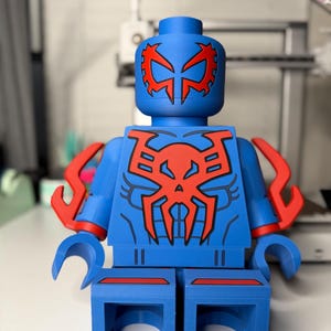 Op de afbeelding: Een grote blauwe figuur die lijkt op een Lego-figuur, met rode accenten. De figuur heeft een rood spinachtig embleem op de borst en het hoofd, met extra rode details op de armen. De figuur staat op een wit oppervlak.