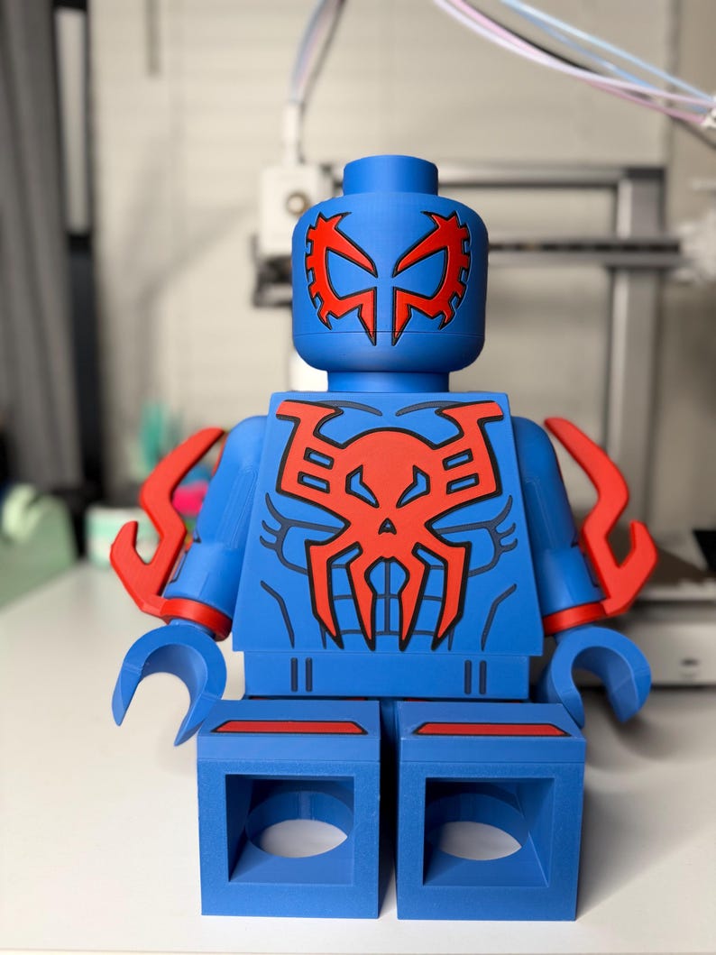 Op de afbeelding: Een grote blauwe figuur die lijkt op een Lego-figuur, met rode accenten. De figuur heeft een rood spinachtig embleem op de borst en het hoofd, met extra rode details op de armen. De figuur staat op een wit oppervlak.