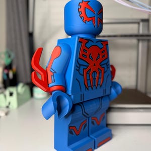 Op de afbeelding: Een grote blauwe figuur die lijkt op een Lego-minifiguur, met rode accenten en een gedetailleerd ontwerp. De figuur heeft een helm met een rood embleem en een spinnenachtig symbool op de borst. De armen en benen hebben rode details. De figuur is ongeveer 30 cm hoog.