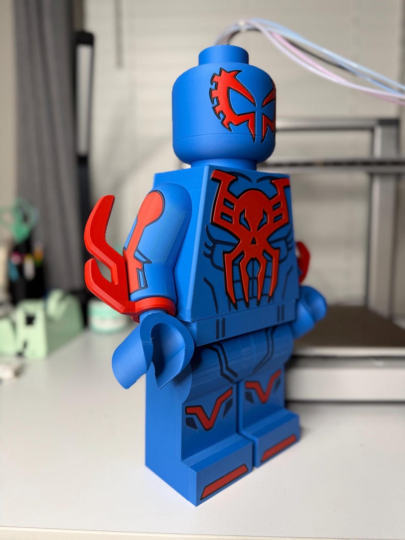 Op de afbeelding: Een grote blauwe figuur die lijkt op een Lego-minifiguur, met rode accenten en een gedetailleerd ontwerp. De figuur heeft een helm met een rood embleem en een spinnenachtig symbool op de borst. De armen en benen hebben rode details. De figuur is ongeveer 30 cm hoog.