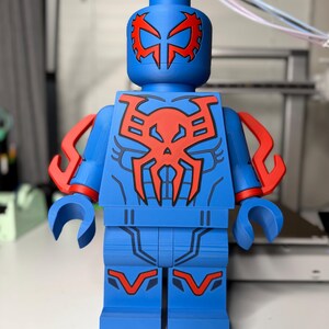 Op de afbeelding: Een grote blauw en rode Lego-achtige figuur. De figuur heeft een blauwe body met rode accenten op het hoofd, de borst en de armen. Het ontwerp van de figuur doet denken aan een superheldenpak. De figuur staat rechtop.