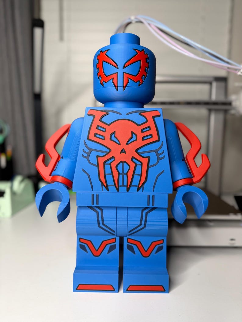 Op de afbeelding: Een grote blauw en rode Lego-achtige figuur. De figuur heeft een blauwe body met rode accenten op het hoofd, de borst en de armen. Het ontwerp van de figuur doet denken aan een superheldenpak. De figuur staat rechtop.
