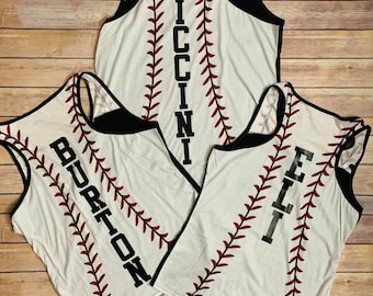 May include: Three white tank tops featuring a red baseball stitch pattern running vertically down the centre. Each tank top displays a black name printed vertically: PICCINI, BURTON, and ELI. The garments have black trim around the armholes and neck.