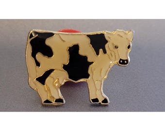 May include: A metal enamel pin shaped like a cow with a cream-colored body and irregular black patches. The cow is depicted in profile facing right, featuring a gold-toned metal outline, black hooves, and a small black nose detail.
