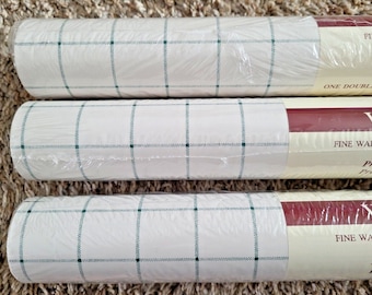 May include: Three rolls of wallpaper wrapped in clear plastic, featuring a white background with a dark green grid pattern. Each roll has a label on the right side with the text FINE WALLCOVERINGS and ONE DOUBLE ROLL, printed in white.