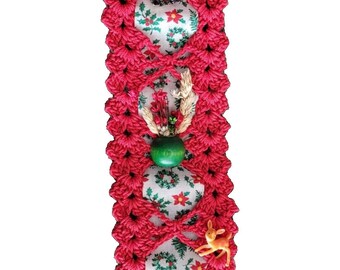 May include: A vertical red crocheted fabric strip featuring a repeating diamond pattern. The strip overlays a white fabric ribbon printed with green holly leaves, red berries, and poinsettias. Embellishments include a Santa figure, a green bead, a small deer, and a child figure.