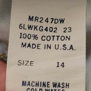 May include: A white tag with black text detailing the size, fabric content, and care instructions for a garment. The tag reads "MR2 47DW 6LWKG402 23 100% COTTON MADE IN U.S.A. SIZE 14 MACHINE WASH COLD WATER LINE DRY WPL 6428".