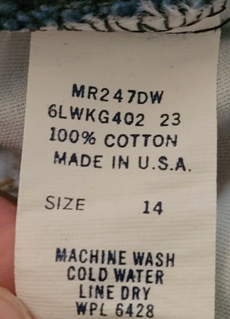 May include: A white tag with black text detailing the size, fabric content, and care instructions for a garment. The tag reads "MR2 47DW 6LWKG402 23 100% COTTON MADE IN U.S.A. SIZE 14 MACHINE WASH COLD WATER LINE DRY WPL 6428".