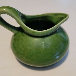 May include: A small, round ceramic creamer pitcher featuring a glossy, variegated green glaze. The pitcher has a curved handle on one side and a pointed pouring spout, with a smooth, rounded body and a deep, open top for holding liquids.