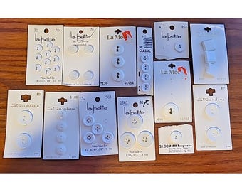 May include: A collection of white buttons packaged on individual white cardboard cards. The cards feature brand names including La Petite, La Mode, and Streamline, with various button counts, sizes like 7/16 inch, and price tags printed on them.