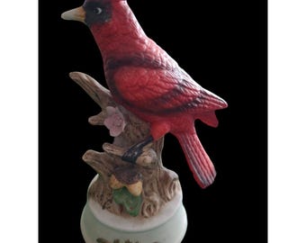 May include: A ceramic figurine of a bright red cardinal bird perched on a textured brown tree branch. The branch features a small pink flower and a green leaf near the base. The bird has a black face mask and a light-colored beak.