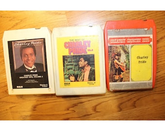May include: Three 8-track tape cartridges featuring Charley Pride. The left is white with a black label, the center is white with a yellow label, and the right is orange with a silver and yellow label. All display text including Greatest Hits.