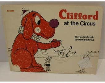 May include: A children's book titled Clifford at the Circus by Norman Bridwell. The cover features a large red dog wearing a white collar and a small hat, holding a trapeze with a person on it, alongside a small child and circus tents.